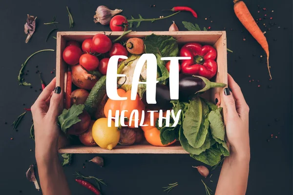 Cropped image of woman holding wooden box with vegetables above gray table with "eat healthy" lettering — Stock Image Cropped Image Woman Holding Wooden Box Vegetables Gray Table Eat Stock Picture