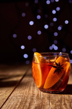 close up view of hot mulled wine drink with orange pieces and anise stars on wooden surface with bokeh lights on background