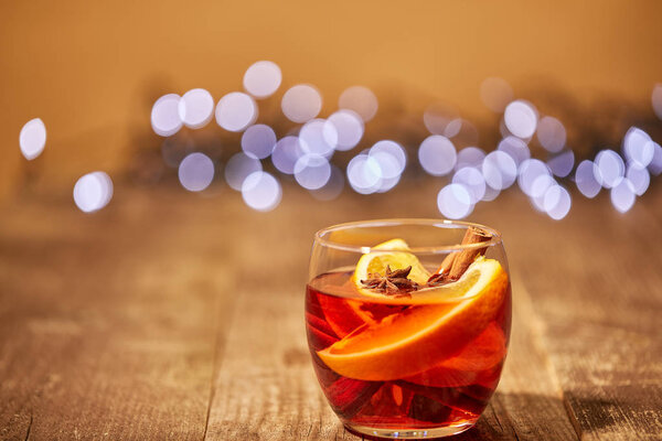 close up view of mulled wine drink with orange pieces and anise stars on wooden surface with bokeh lights on background