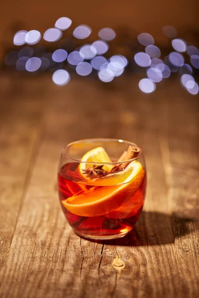 close up view of mulled wine drink with orange pieces and anise stars on wooden surface with bokeh lights on background