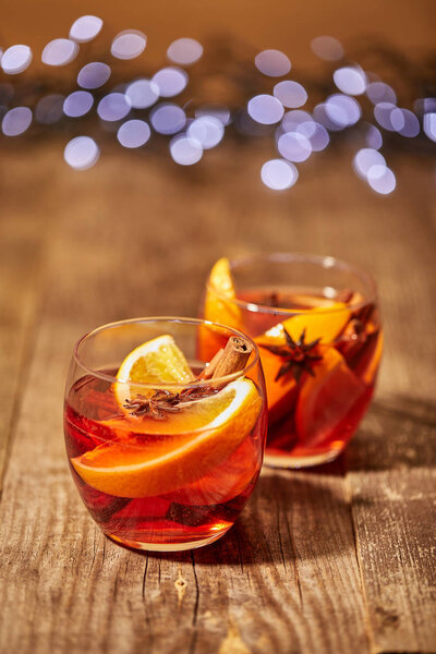 close up view of mulled wine in glasses with orange pieces and spices on wooden surface with bokeh lights on backdrop