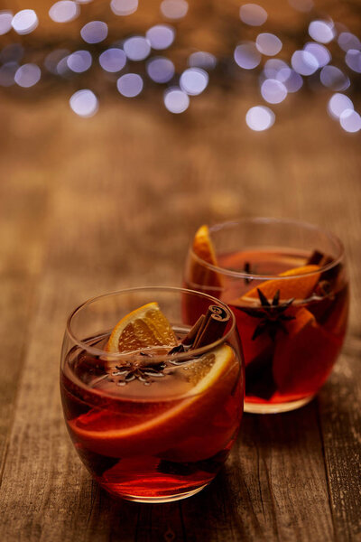 close up view of delicious hot mulled wine in glasses with orange pieces on wooden tabletop with bokeh lights on backdrop