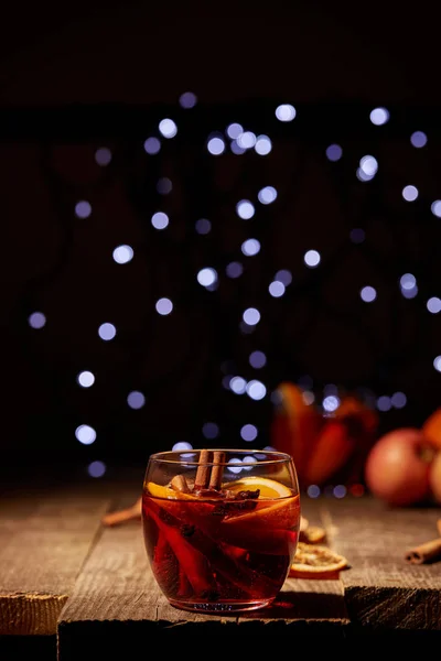 close up view of hot mulled wine drink with orange pieces and anise stars on wooden surface with bokeh lights on background