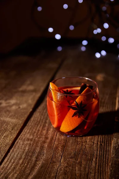 close up view of hot mulled wine drink with orange pieces and anise stars on wooden surface with bokeh lights on background
