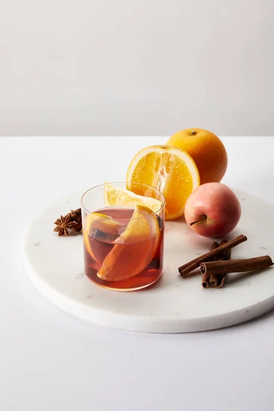 close up view of hot mulled wine in glass fresh oranges and apple on white surface on grey backdrop