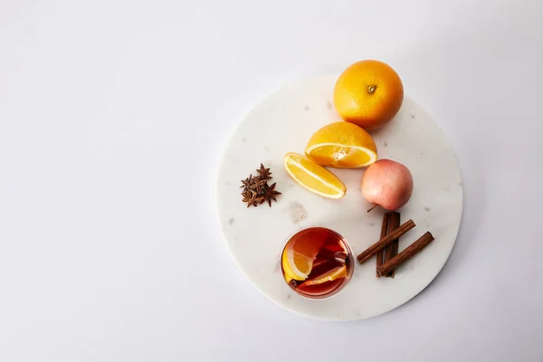 top view of hot mulled wine in glass, spices, oranges and apple on white surface