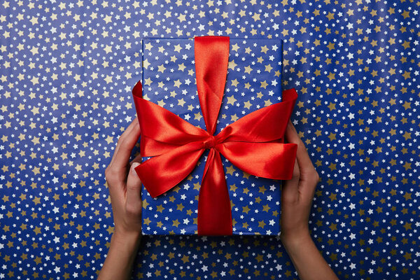 cropped shot of woman holding bright wrapped gift with red ribbon on wrapping paper with stars pattern