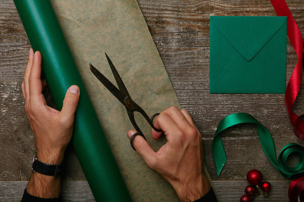 частичный вид человека с ножницами, оберточная бумага для упаковки подарков и конверт на деревянном столе
