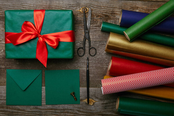 плоский с рождественским подарком с красной лентой, оберточной бумагой, ножницами и конвертами для поздравительной открытки на деревянной поверхности
