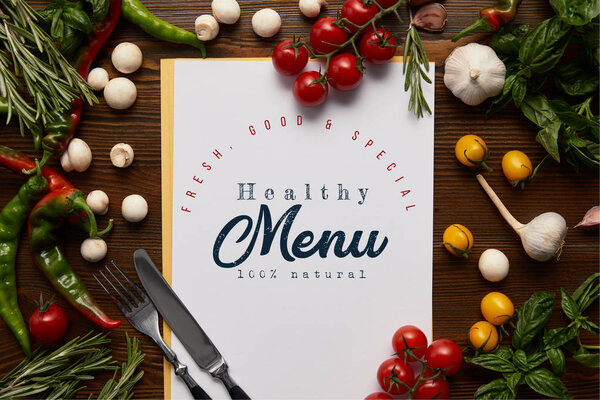 top view of card with "healthy menu" lettering, cutlery and fresh herbs with vegetables on wooden surface 