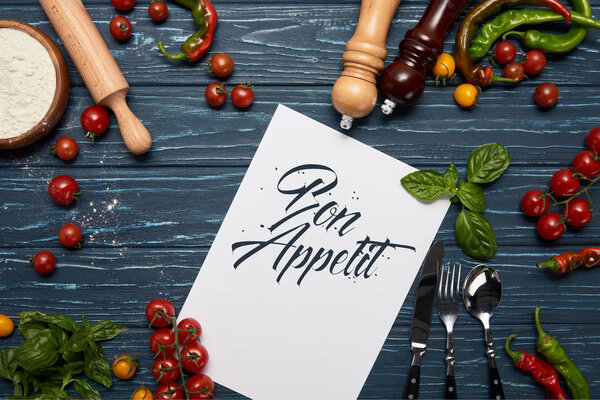 top view of template with "bon appetit" lettering, fresh vegetables, cutlery and spices on wooden surface