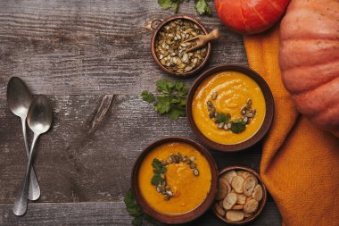 top view of delicious pumpkin soup in bowls, spoons, fresh pumpkins and rusks on wooden table clipart