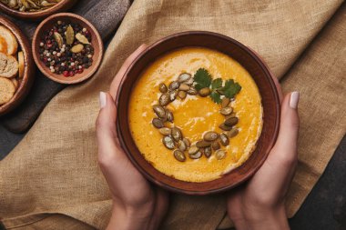 partial top view of person holding bowl with tasty pumpkin soup above sackcloth clipart