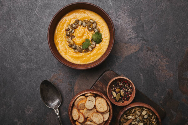 bowl with tasty pumpkin soup, spices, rusks, pumpkin seeds and spoon on dark surface 