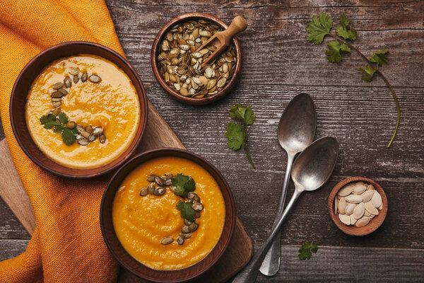 top view of bowls with pumpkin soup, spoons and pumpkin seeds on rustic wooden table