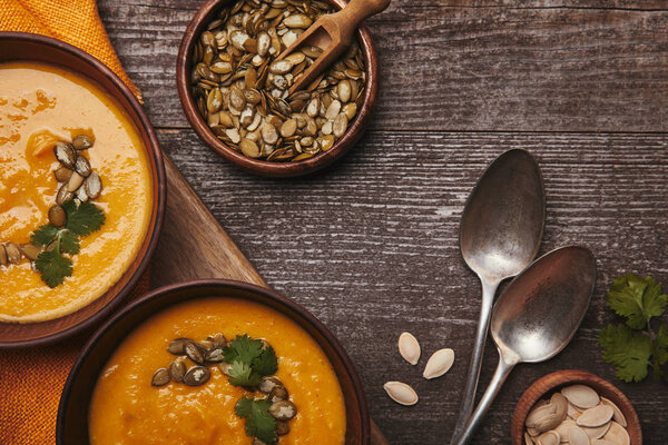 top view of tasty healthy pumpkin soup in bowls, spoons and pumpkin seeds on wooden table