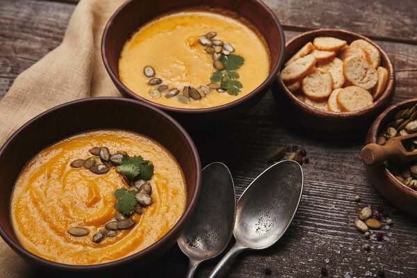 bowls with pumpkin soup, spoons, pumpkin seeds and rusks on wooden table