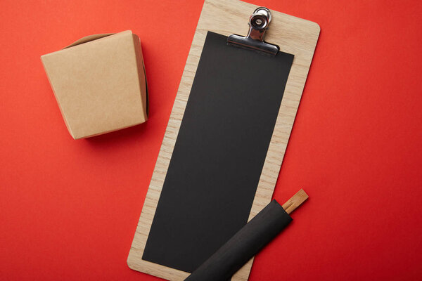 flat lay with noodle box, chopsticks and blank black menu on red surface