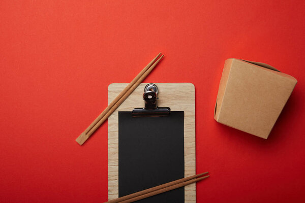 view from above of arranged noodle box, chopsticks and blank black menu on red surface