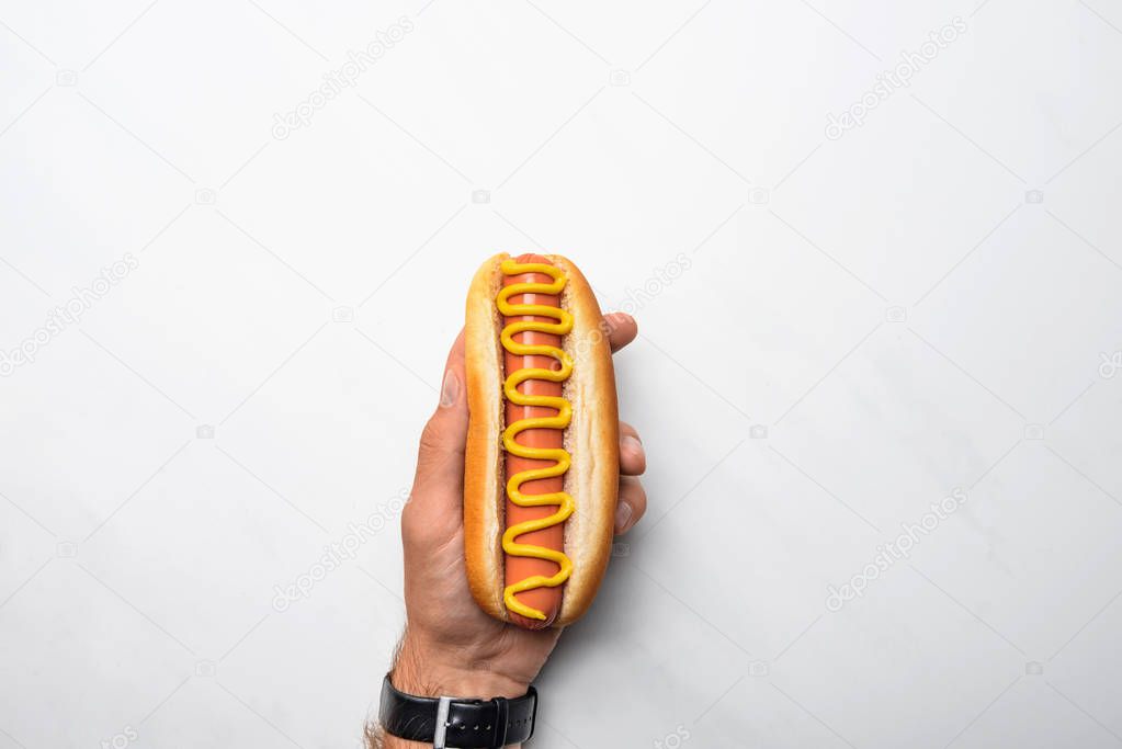 cropped shot of man holding tasty hot dog poured with mustard on white marble surface