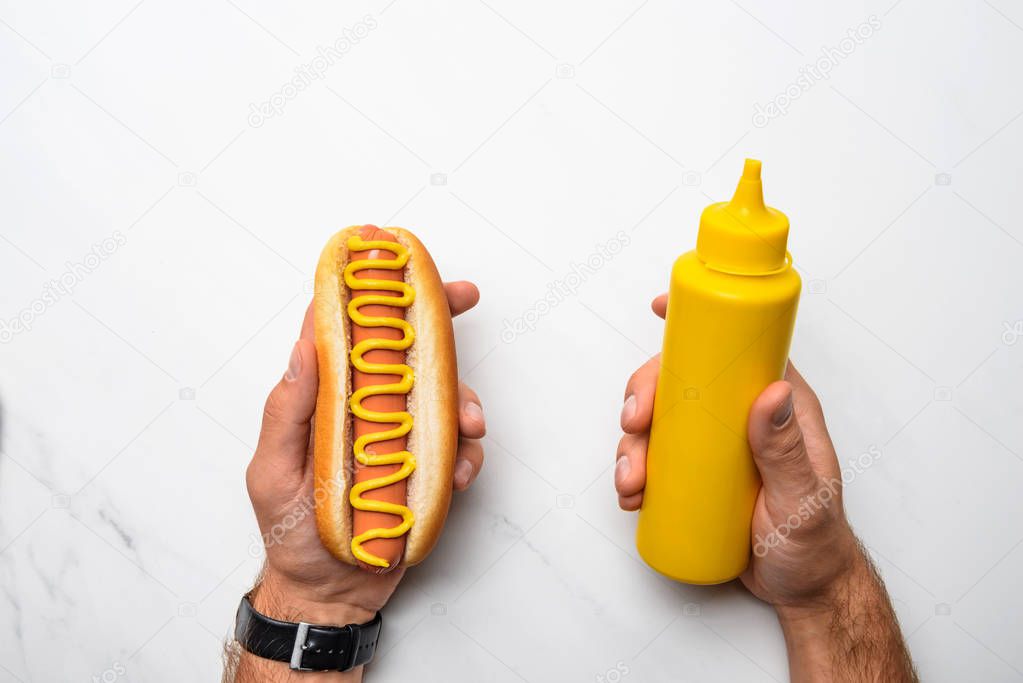 cropped shot of man holding tasty hot dog with mustard on white marble surface