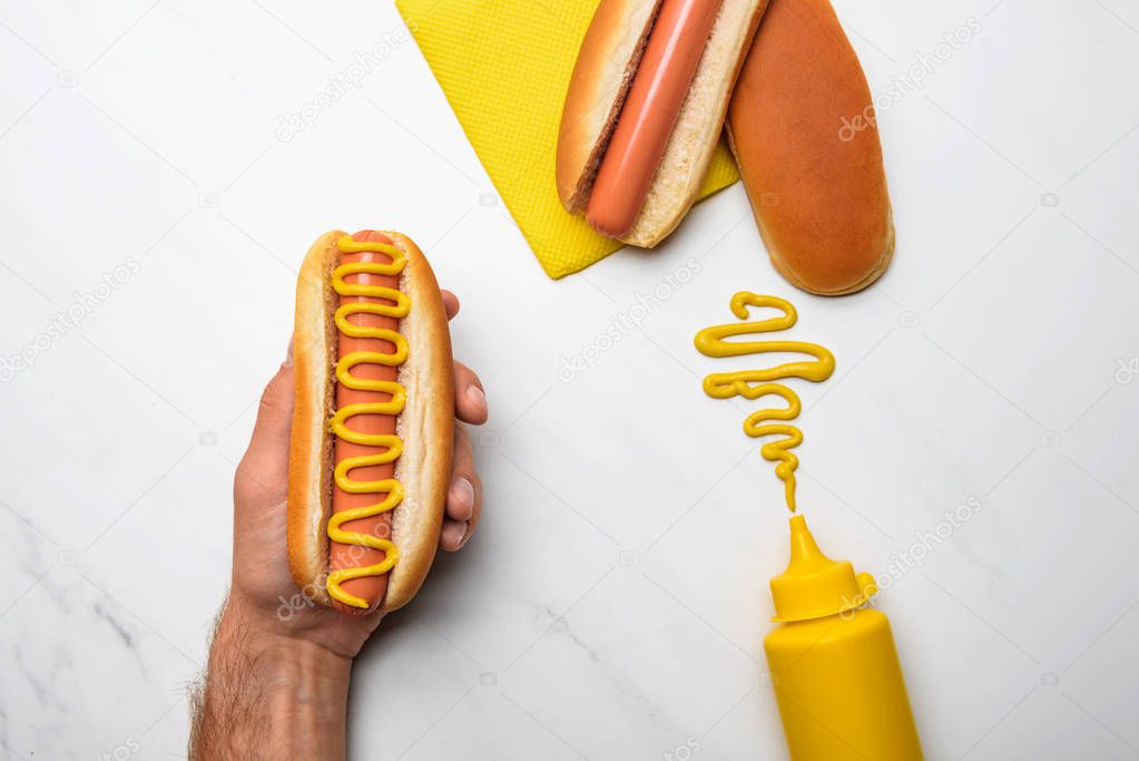 cropped shot of man holding hot dog with mustard on white marble surface