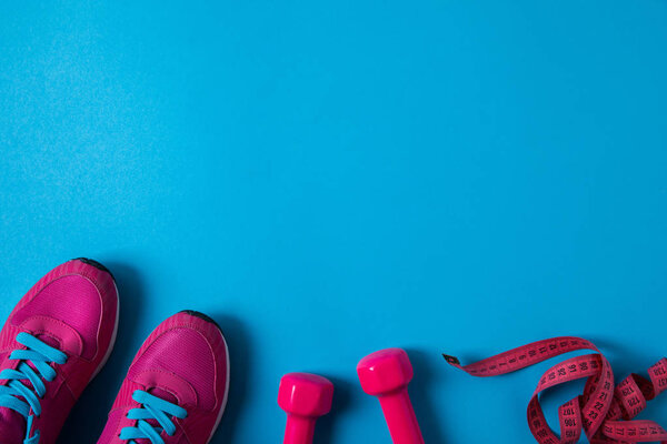 elevated view of pink sneakers, dumbbells and measuring tape placed in row isolated on blue