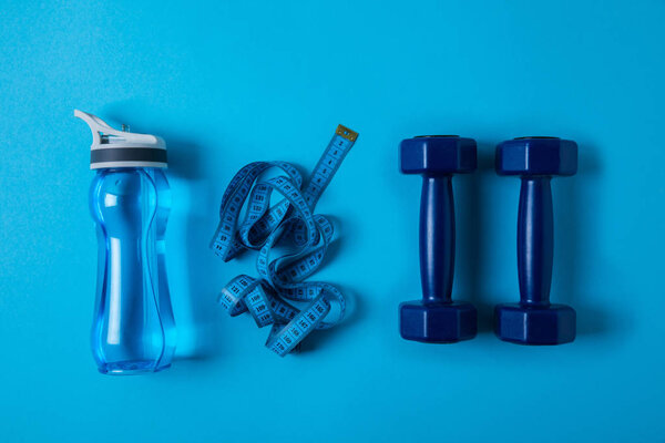 view from above of sport bottle with water, measuring tape and dumbbells isolated on blue 