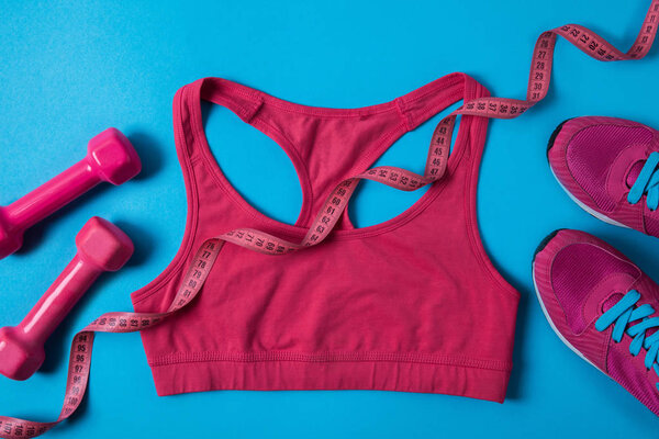 top view of pink dumbbells, measuring tape, sports bra and sneakers isolated on blue 
