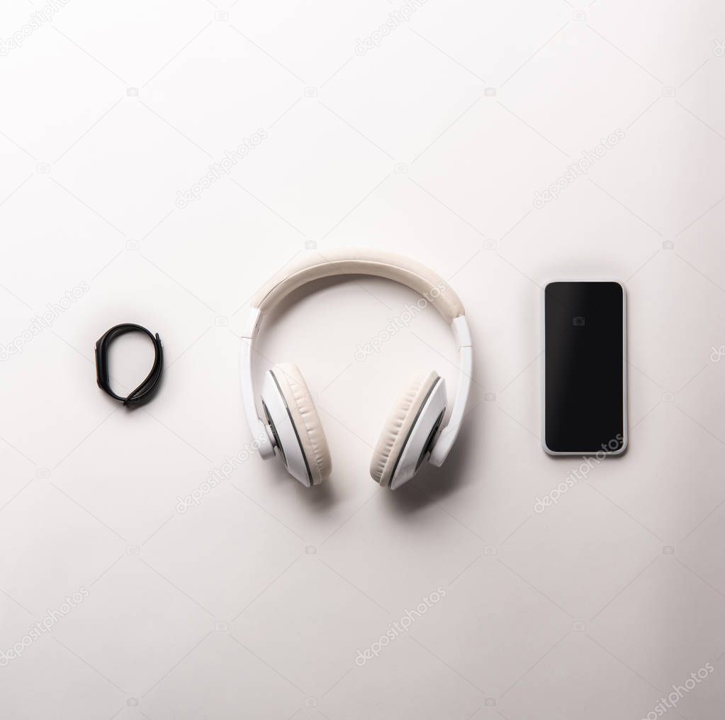 top view of fitness tracker, headphones and smartphone with blank screen placed in row isolated on white, minimalistic concept 