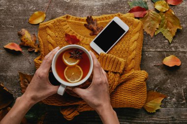 kısmi görünümünü çay örme kazak, smartphone ve düşen yapraklar ile ahşap masa üzerinde tutan kadın