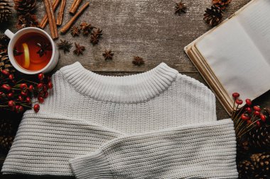 flat lay with white sweater, red holly berries, cinnamon sticks, blank notebook and cup of tea on wooden surface