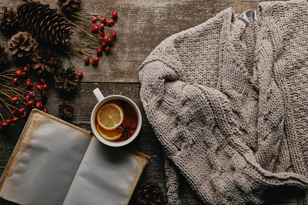 flat lay with red holly berries, blank notebook, cup of tea and knitted sweater on wooden tabletop