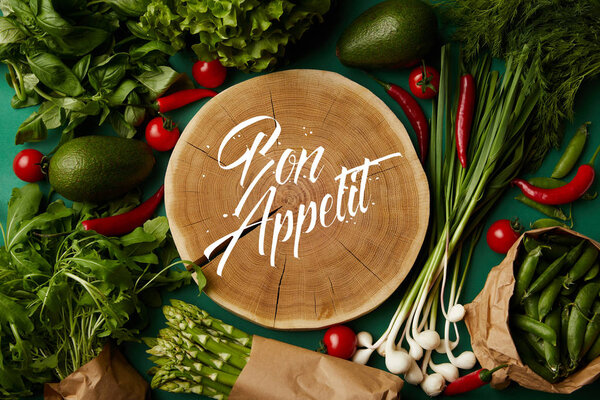 top view of wood cut with bon appetit inscription surrounded with different ripe vegetables on green surface