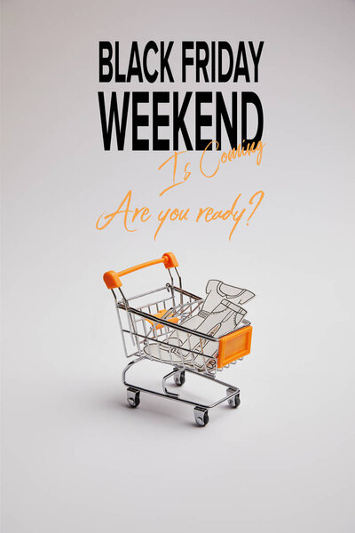 close up view of shopping cart with little goods made of paper on grey background, black friday weekend inscription