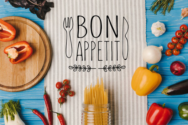 top view of uncooked pasta and ripe vegetables on napkin on blue table, bon appetit lettering