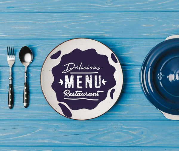 Top view of fork, spoon and plates on blue table, delicious menu restaurant lettering — Stock Image Top View Fork Spoon Plates Blue Table Delicious Menu Restaurant Stock Picture