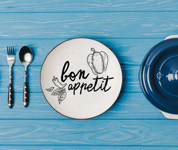 Top view of fork, spoon and plates on blue table, bon appetit lettering — Stock Image Top View Fork Spoon Plates Blue Table Bon Appetit Lettering Stock Picture