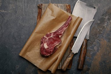 top view of raw rib eye steak on baking paper with kitchen knives on grey background clipart