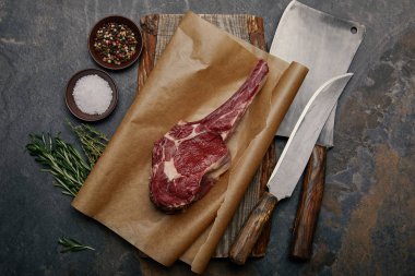top view of raw rib eye steak on baking paper with kitchen knives, spices, herbs and salt on grey background clipart