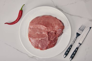 top view of fresh raw meat on plate with kitchen cutlery and chilli pepper on white background clipart