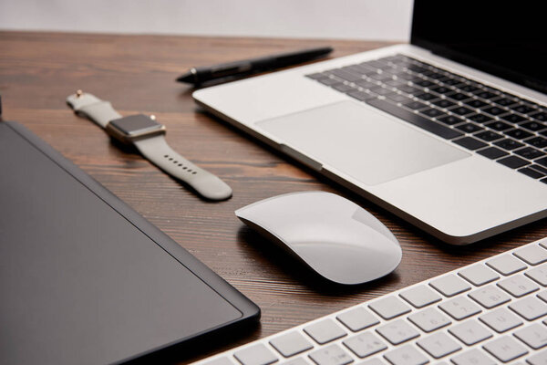 close-up shot of laptop with wireless mouse and keyboard lying on wooden table with smart watch and graphics tablet