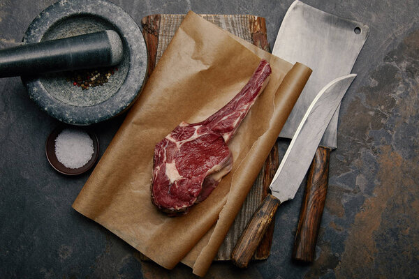 top view of raw rib eye steak on baking paper with kitchen knives and marble pounder on grey background