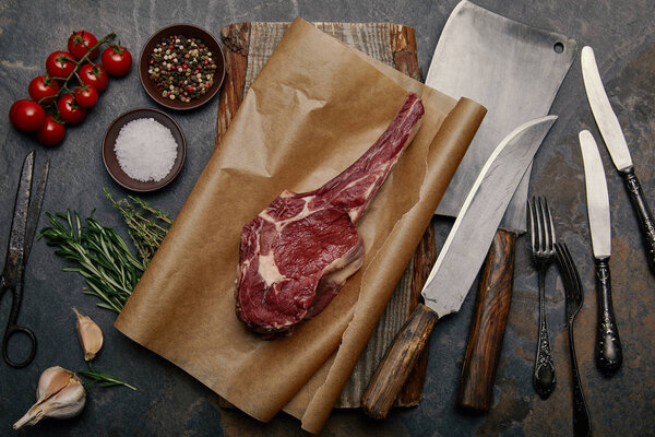 top view of raw rib eye steak on baking paper with variety of kitchen knives, spices, herbs and tomatoes on grey background