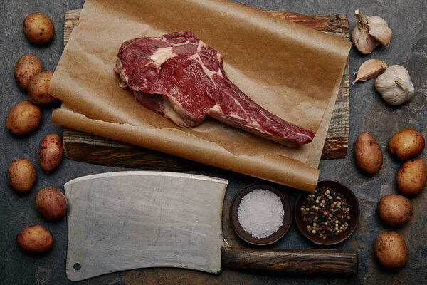 flat lay with raw rib eye steak on baking paper with butcher knife, spices, potatoes and garlic