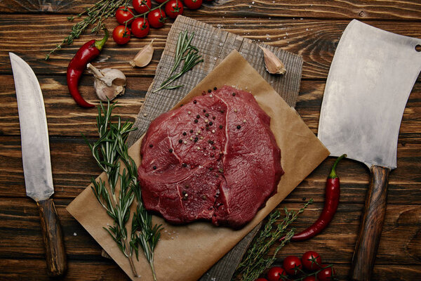 flat lay with raw meat on baking paper with kitchen knives and vegetables on wooden background