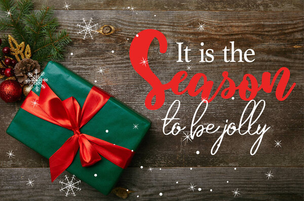 верхний вид завернутый зеленый подарок с красной лентой и рождественские игрушки на деревянном фоне с "это сезон, чтобы быть веселым" вдохновение
