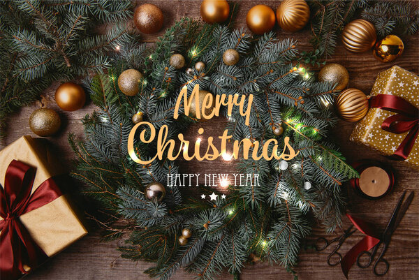 top view of Christmas gift boxes and fir wreath on wooden table with "merry christmas and happy new year" lettering and glowing lights