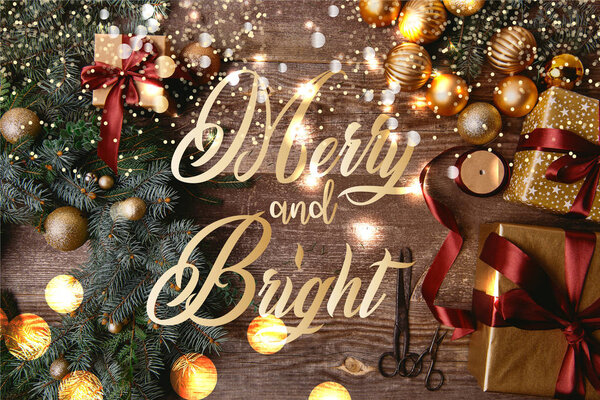 elevated view of Christmas gift boxes, fir wreath, ribbon spool and scissors on wooden table with "merry and bright" lettering and glowing lights