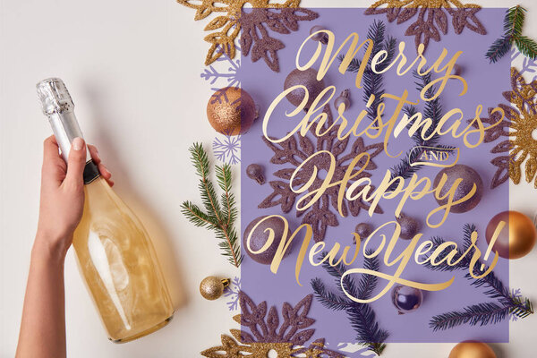 cropped image of woman holding bottle of sparkling champagne near christmas decoration on white with "merry christmas and happy new year" lettering